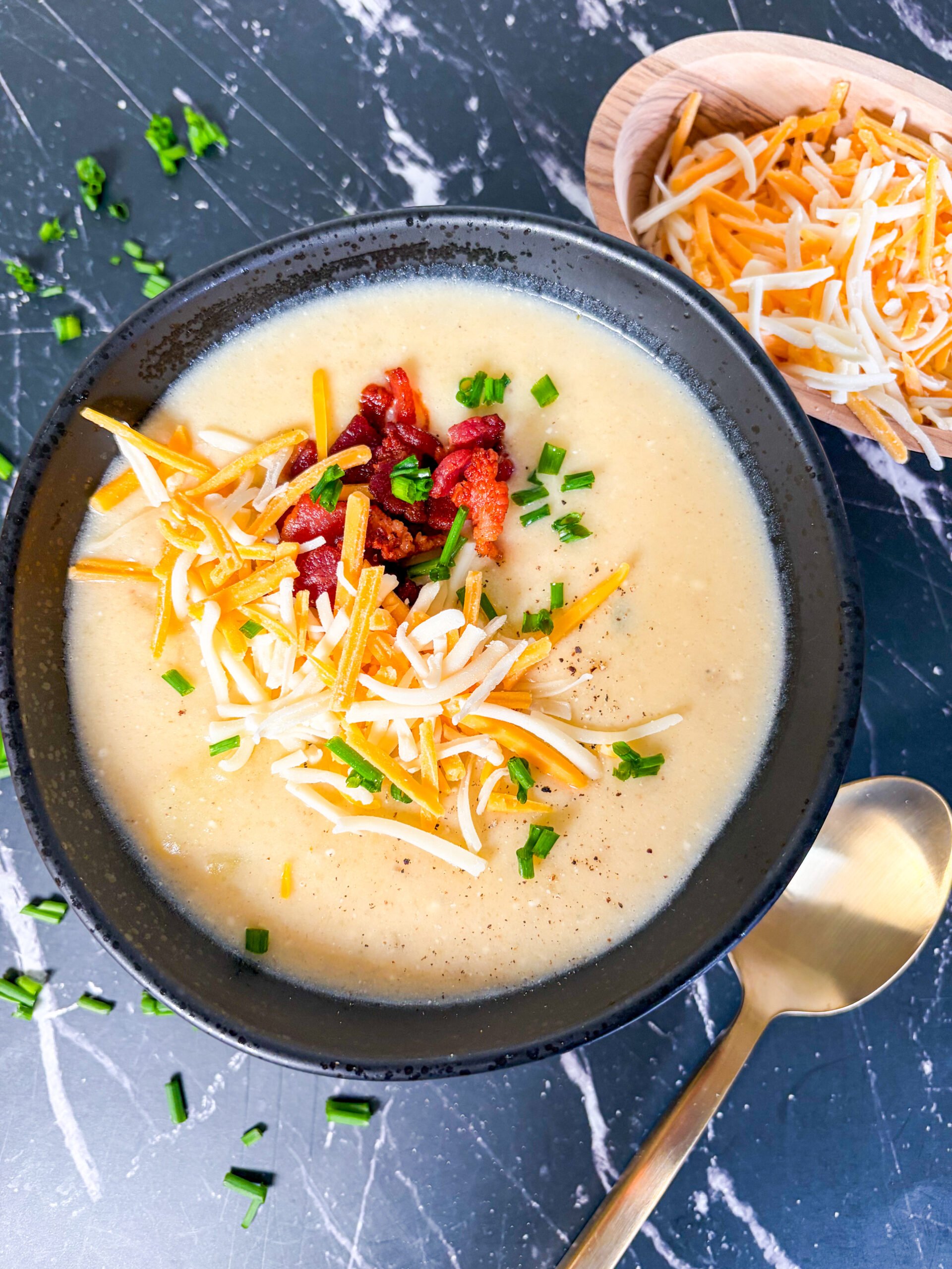 Potato soup in a black bowl topped with cheese, bacon and chive