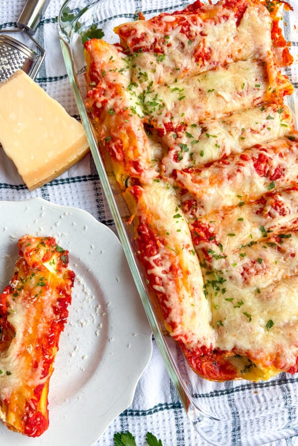 Manicotti in a baking dish and one on a plate next to parm block