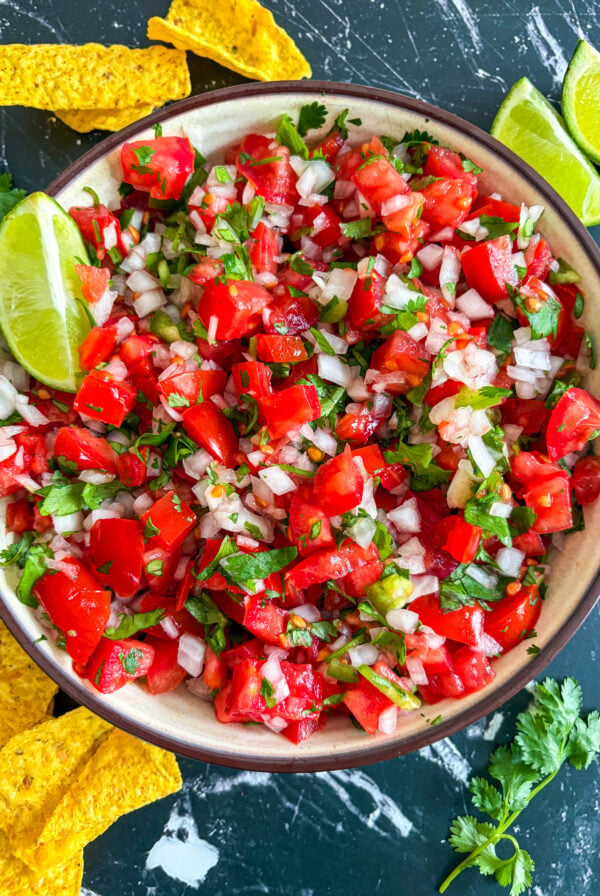 Pico de gallo in a bowl next to tortillas chips, lime wedges, and cilantro