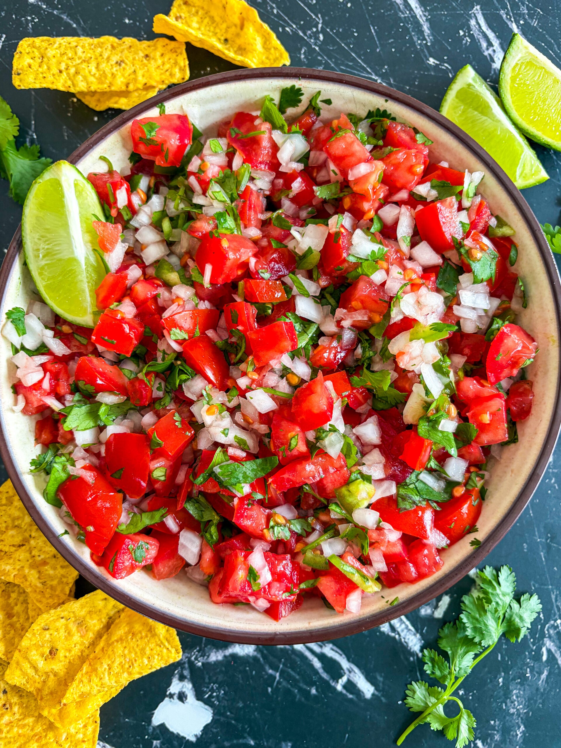 Pico de gallo in a bowl next to tortillas chips, lime wedges, and cilantro