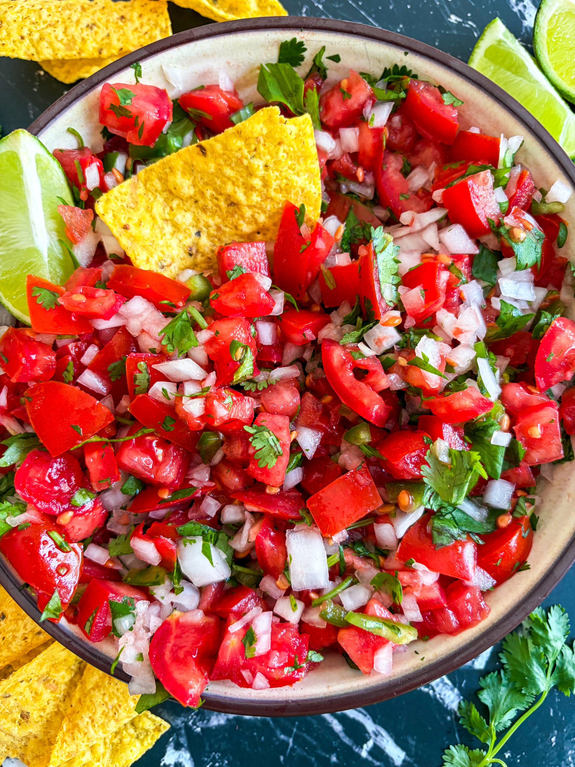 Close up of pico de gallo in bowl next to tortillas chips, lime wedges and cilantro