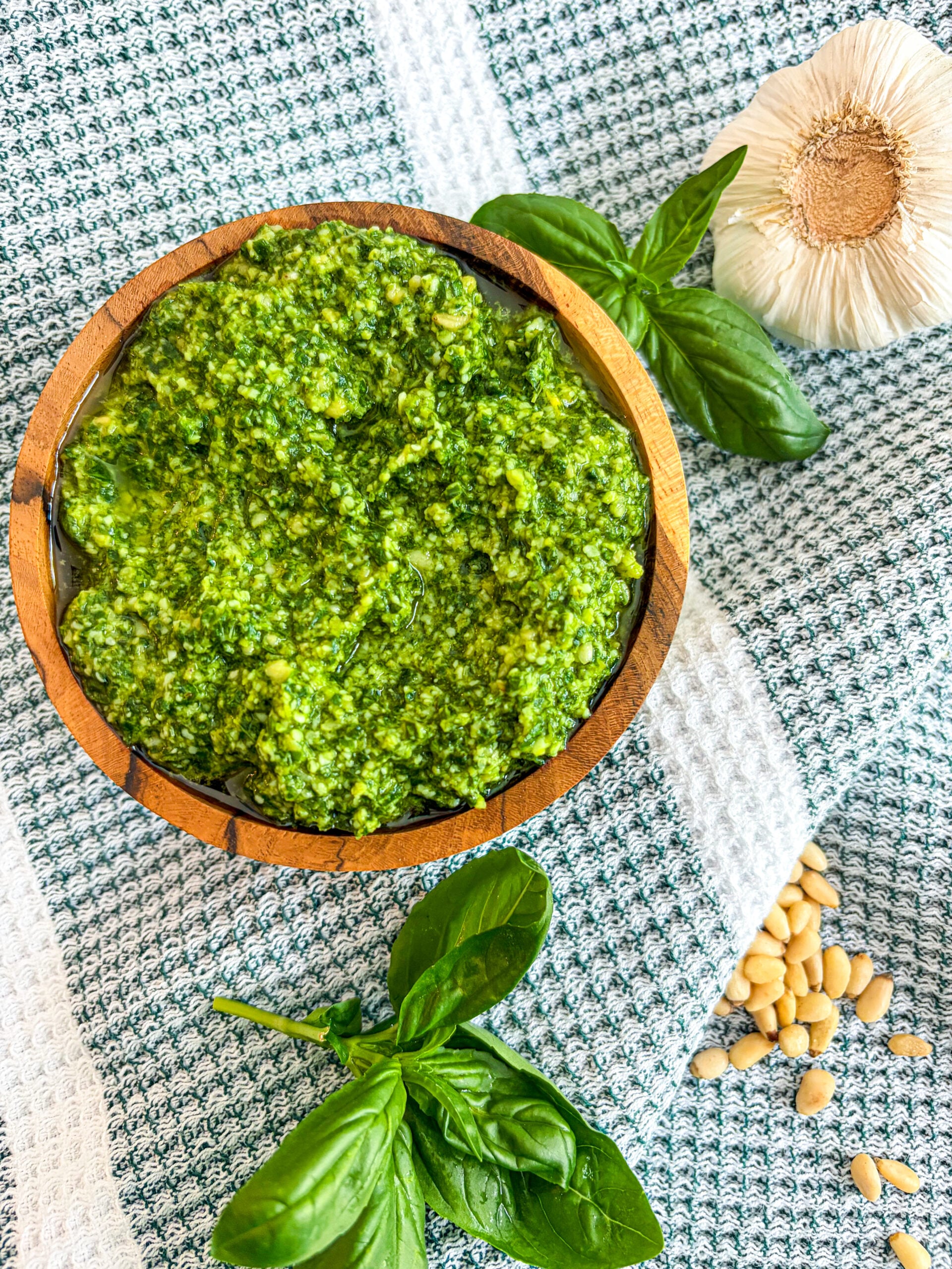 Pesto in a wooden bowl with spoon next to basil, garlic head, and pine nuts