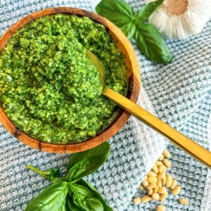 Close up of pesto in a wooden bowl with spoon next to basil, garlic head, and pine nuts