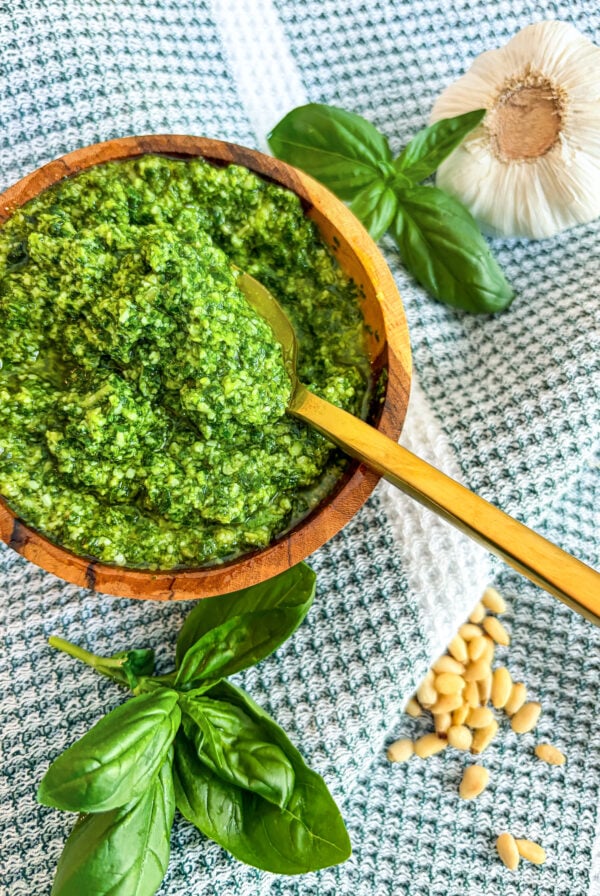 Close up of pesto in a wooden bowl with spoon next to basil, garlic head, and pine nuts