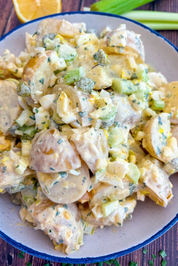 Potato salad in a blue and white serving bowl next to lemon wedge, celery sticks and snipped chives