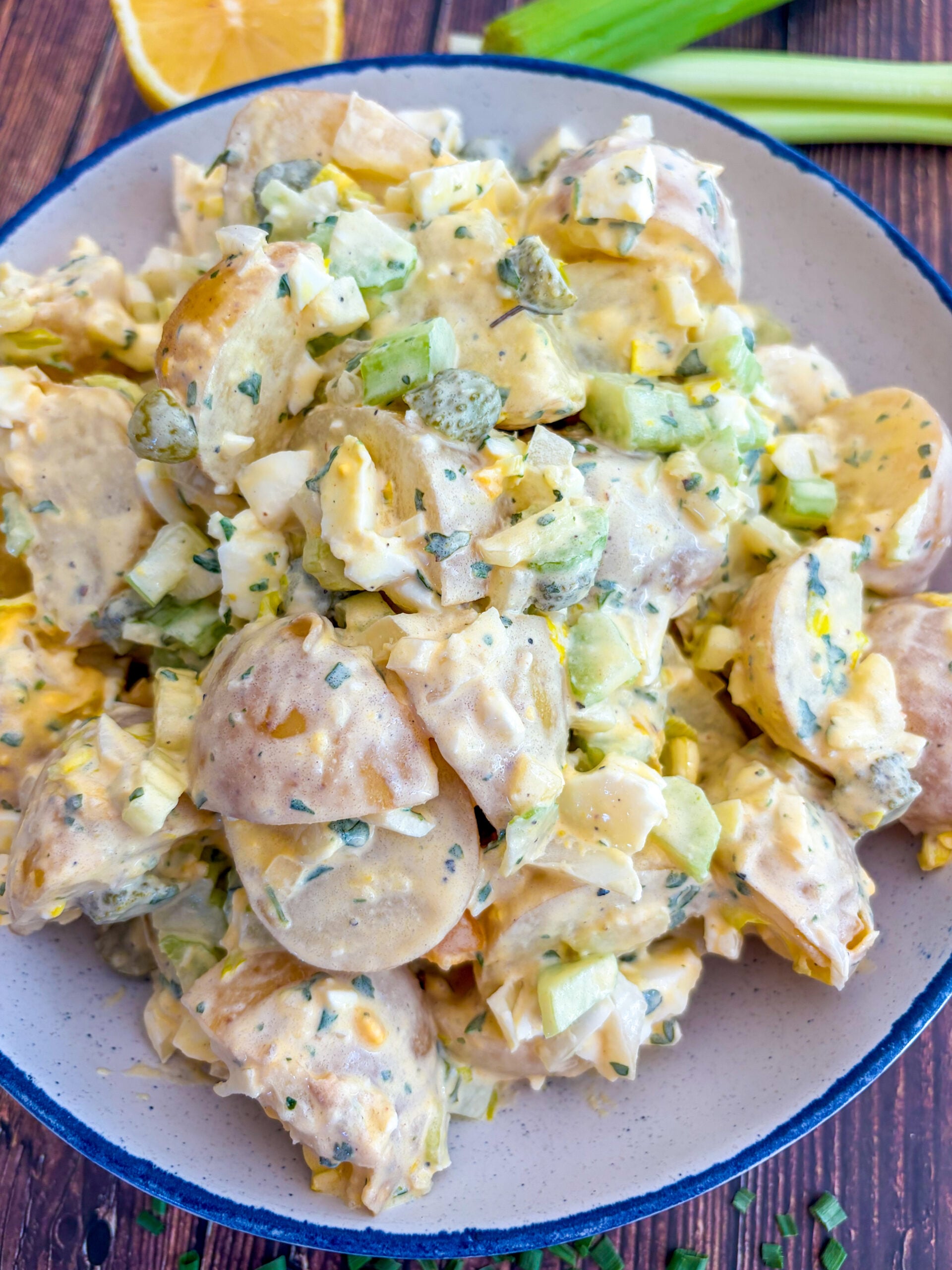 Potato salad in a blue and white serving bowl next to lemon wedge, celery sticks and snipped chives