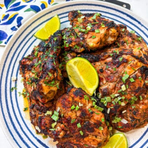 Jerk chicken garnished with scallion and lime wedges on a blue and white plate next to a napkin, fork and knife