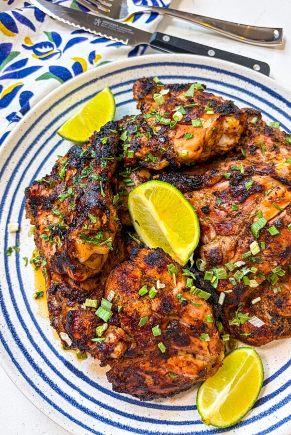 Jerk chicken garnished with scallion and lime wedges on a blue and white plate next to a napkin, fork and knife