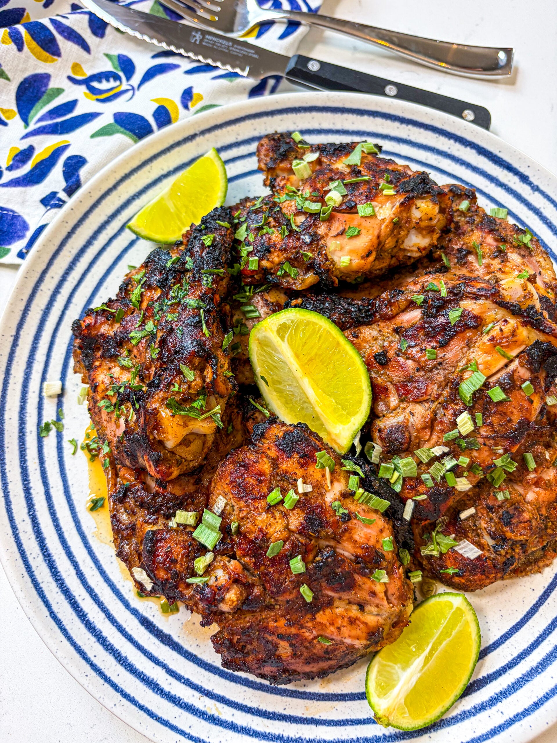 Jerk chicken garnished with scallion and lime wedges on a blue and white plate next to a napkin, fork and knife