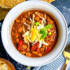 Chicken chili in a white bowl next to spoon, scallions, cheese, and chips