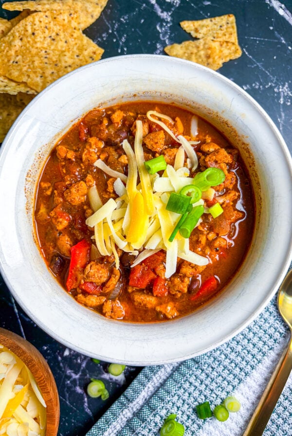 Chicken chili in a white bowl next to spoon, scallions, cheese, and chips