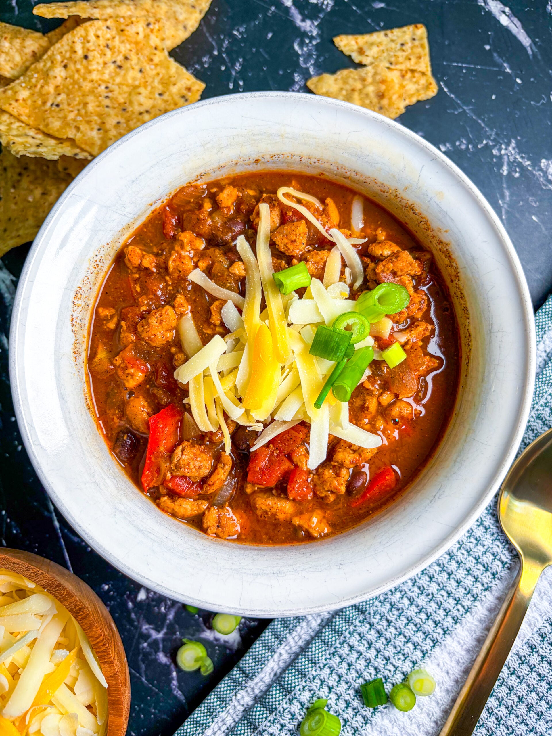 Chicken chili in a white bowl next to spoon, scallions, cheese, and chips