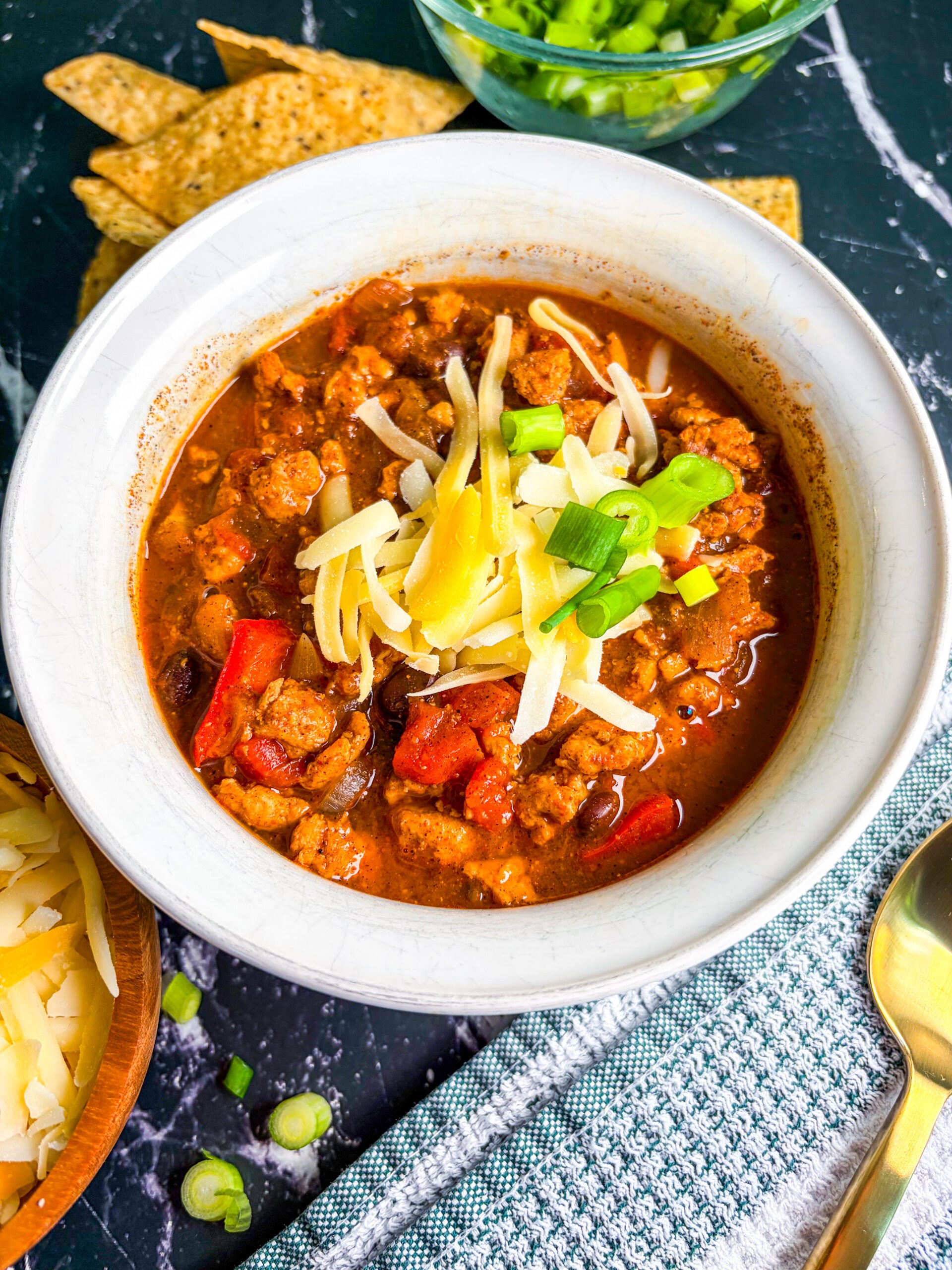 Side view chicken chili in white bowl next to chips, scallions, cheese and spoon
