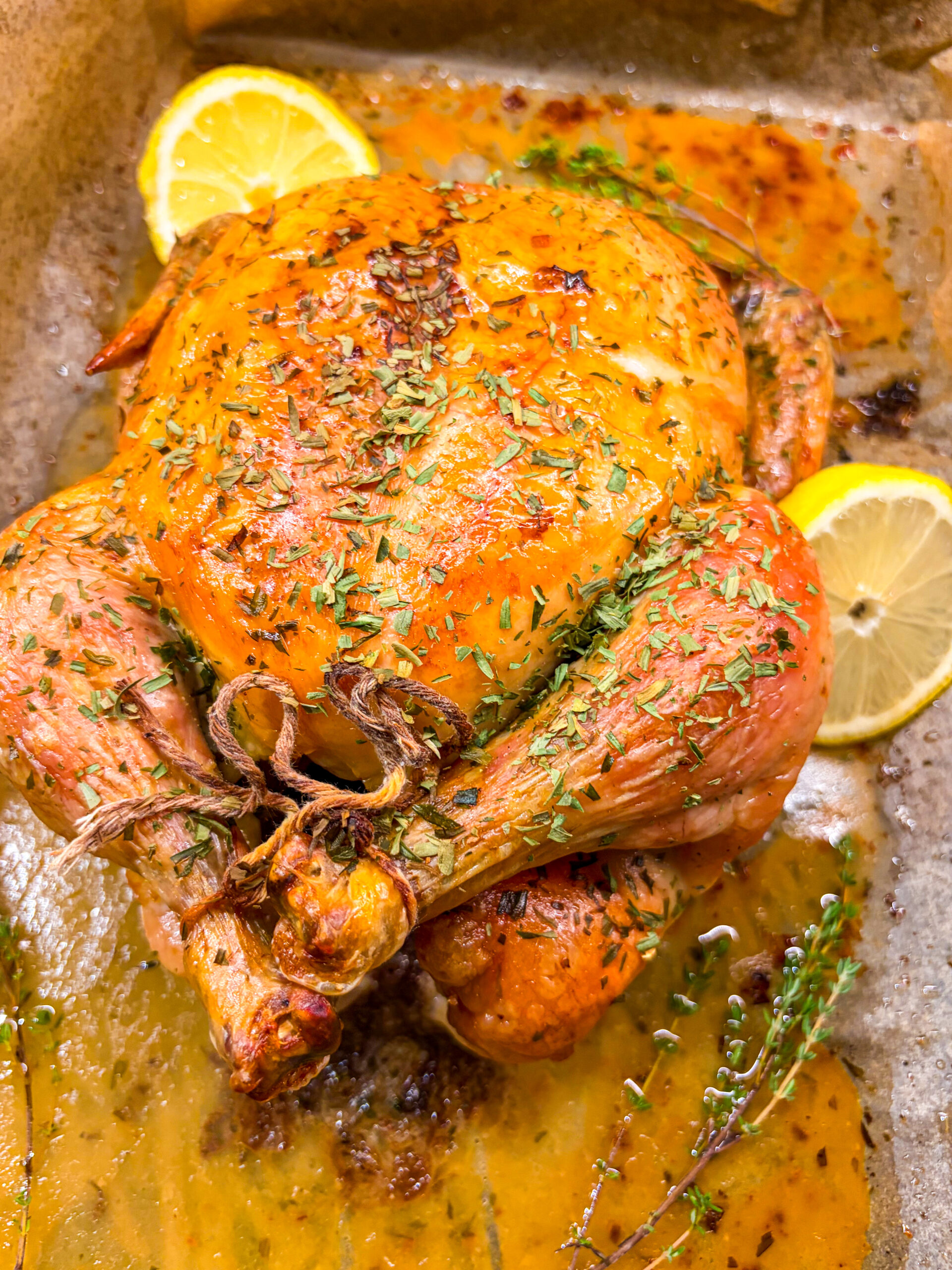 Roast chicken in a roasting pan next to herbs and lemon slices